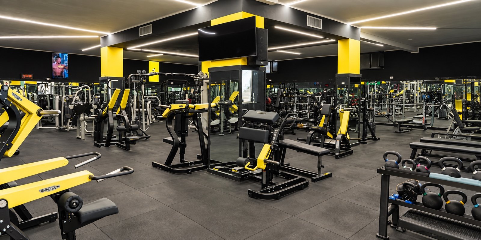 Vista panoramica della sala pesi Athena Wellness, con macchine gialle e nere, specchi e kettlebell